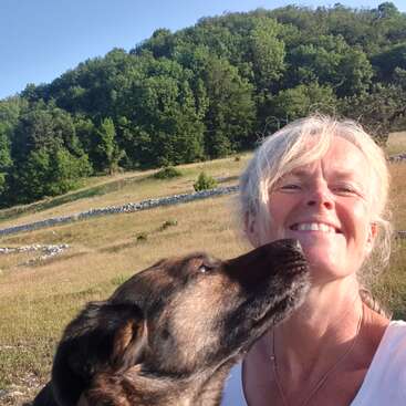 Uma mulher sorridente tira uma selfie ao ar livre com um cachorro marrom lambendo sua bochecha. Eles estão rodeados por campos gramados e árvores verdes sob um céu azul claro.