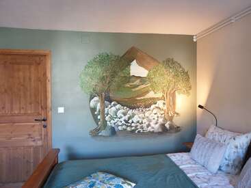 Este quarto aconchegante tem um mural inspirado na natureza, com duas árvores frondosas, um rio rochoso e montanhas. A porta de madeira, a roupa de cama verde e as almofadas com estampas suaves completam o visual.