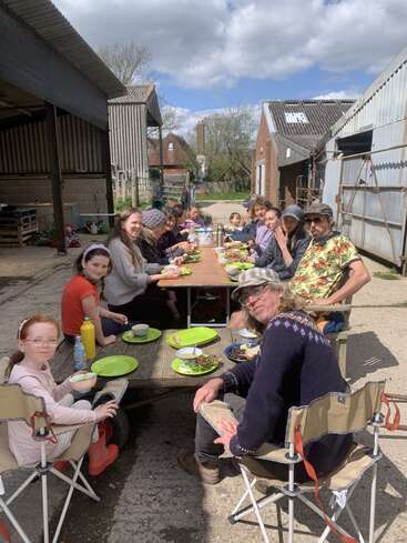 La imagen muestra a un grupo de personas, niños y adultos, reunidos en torno a una larga mesa al aire libre, disfrutando de una comida en un día soleado.