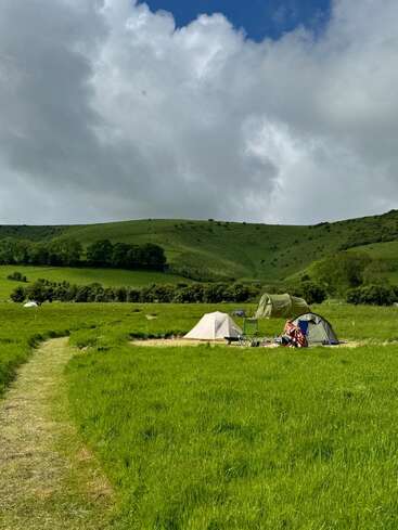 La imagen representa un sereno campo de hierba con un camino serpenteante, en el que aparecen tres tiendas y una silla, con un telón de fondo de colinas ondulantes y un cielo nublado.