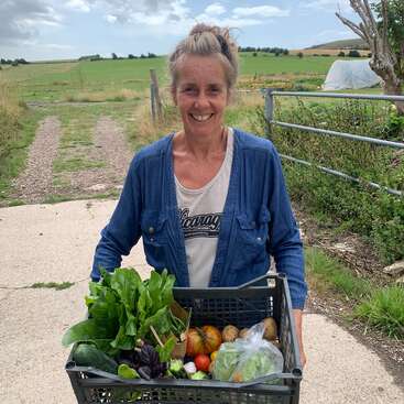 Una mujer de pelo rubio, con camisa azul y camiseta blanca, lleva una caja negra llena de verduras por un camino en una zona rural.