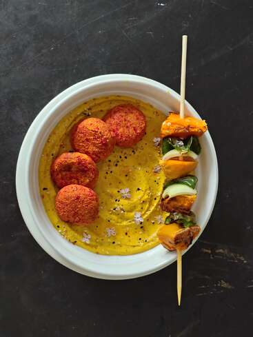 Um prato branco contém quatro bolinhos de falafel sobre um molho amarelo cremoso, polvilhados com sementes e decorados com pequenas flores, acompanhados por um espeto de legumes.
