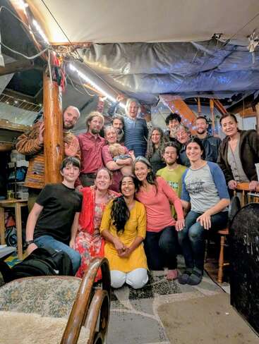 Um grupo alegre de pessoas de várias idades se reúne dentro de casa, sorrindo calorosamente para a câmera. A sala aconchegante está decorada com luzes de corda, criando um clima festivo.