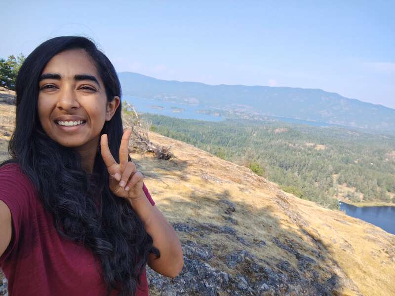 Uma mulher de cabelos longos e escuros sorri e faz o sinal da paz, tirando uma selfie no topo de uma colina, com montanhas, árvores e lagos pitorescos ao fundo.