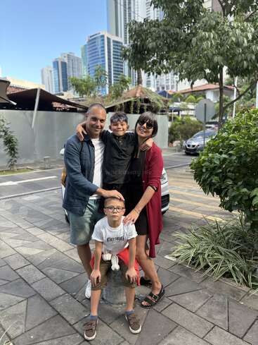 Una familia de cuatro miembros posa en una acera de la ciudad, con edificios altos al fondo. Ambos padres están detrás de sus dos hijos, sonriendo cálidamente juntos.