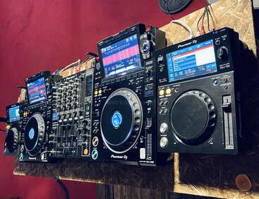 Esta imagem mostra um equipamento profissional de DJ com controladores e mixers da Pioneer DJ com telas grandes, jog wheels, vários botões e controles deslizantes, colocados em uma mesa de madeira.