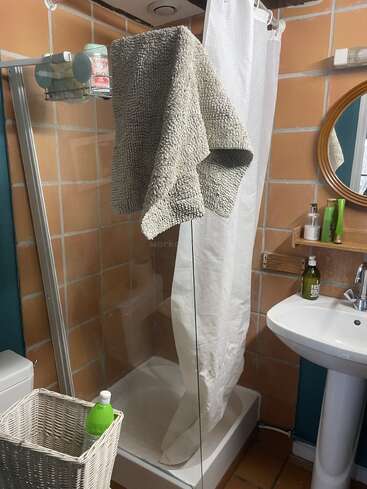 Un acogedor cuarto de baño con azulejos color melocotón cuenta con una cabina de ducha, una cortina blanca, una mullida toalla gris colgada, una cesta de mimbre, una botella verde, un lavabo, un espejo y artículos de tocador en las estanterías.