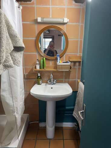 Este acogedor cuarto de baño cuenta con un lavabo con pedestal, un espejo redondo, paredes alicatadas, una ducha con cortina, toallas, una estantería con artículos de aseo y una persona haciéndose un selfi frente al espejo.