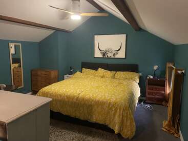 Esta acogedora habitación tiene paredes de color verde azulado, vigas vistas, una cama grande con ropa de cama amarilla, muebles de madera, dos espejos, lámparas de noche y un cuadro con un toro enmarcado.