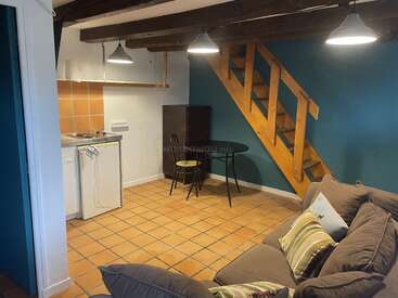 Este acogedor espacio cuenta con baldosas de terracota, vigas de madera, una pequeña cocina, una mesa de comedor redonda, sillas de metal, una escalera, paredes de color verde azulado y un sofá acolchado.