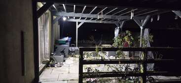 Un patio faiblement éclairé la nuit présente une pergola, des meubles de jardin, des objets épars, des plantes en pot et une clôture en bois avec des plantes grimpantes qui se découpent sur l'obscurité.