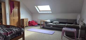 Cette chambre mansardée confortable comprend un lit, un canapé d'angle gris, un tapis de yoga, des poufs, une table rose, un ventilateur, une lucarne, des meubles en bois et un décor neutre.