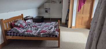 Une chambre mansardée confortable avec un lit en bois, une literie à carreaux, des tables d'appoint, des étagères de placard ouvertes, une moquette beige, une porte en bois et la lumière du soleil filtrant à travers des rideaux à fleurs.