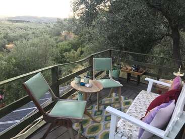 A cozy wooden balcony overlooks a lush green landscape. Two chairs, a small table, and a bench with colorful pillows create a peaceful outdoor retreat.