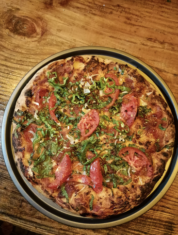 A rustic homemade pizza in a metal pan sits on a wooden table. Topped with fresh tomato slices, green peppers, cheese, herbs, and vibrant greens.