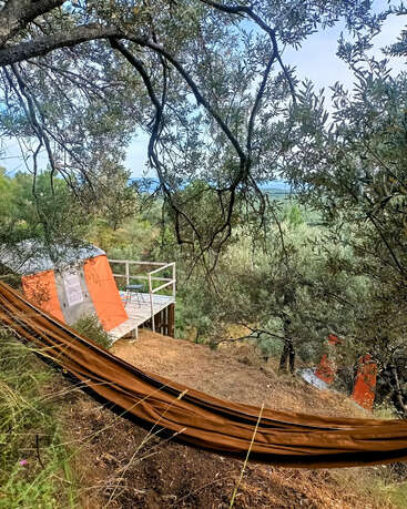 Una acogedora tienda de glamping, con un exterior en tonos naranja y gris, se alza sobre una terraza de madera en medio de frondosos árboles, desde donde se disfruta de unas vistas tranquilas y pintorescas de las verdes colinas.