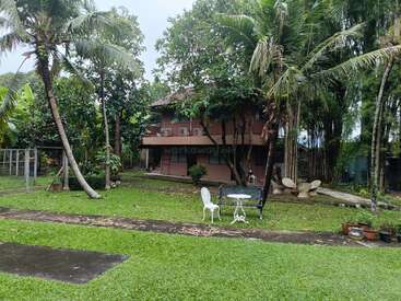 Ein zweistöckiges braunes Haus, umgeben von üppigem Grün, Palmen und einem gepflegten Rasen. Weiße Stühle und ein Tisch im Freien sorgen für eine gemütliche Atmosphäre. Friedliche Umgebung.