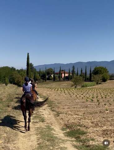 Dos personas cabalgan por un camino de tierra a través de un campo rural, en dirección a una villa rodeada de árboles, con montañas visibles bajo un cielo azul despejado.