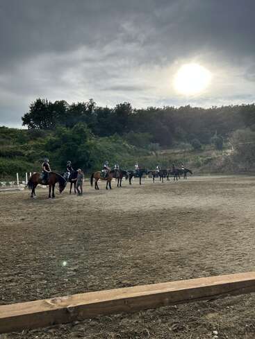 Bajo un cielo nublado, varias personas montan a caballo en un picadero vallado, alineadas una al lado de la otra, con los árboles y el sol de fondo.