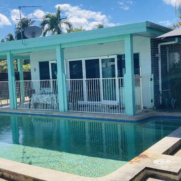 Una piscina cristalina se encuentra frente a una casa moderna con columnas de color turquesa, una valla blanca, puertas correderas y palmeras tropicales bajo un cielo azul y soleado.