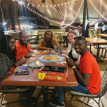 Cinco amigos se sentam em torno de uma mesa em um café aconchegante, sorrindo e jogando o jogo de tabuleiro Catan, cercados por luzes quentes e um ambiente descontraído.
