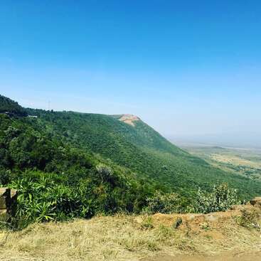 Uma encosta verde exuberante se estende sob um céu azul claro, com grama seca em primeiro plano e uma vasta planície visível no fundo distante.