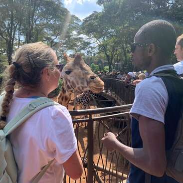 Duas pessoas ficam em pé ao lado de uma cerca, observando uma girafa em um parque de vida selvagem. O dia ensolarado, as árvores e os outros visitantes criam uma atmosfera animada ao ar livre.