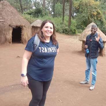 Uma mulher sorridente em roupas casuais fica ao ar livre perto de cabanas tradicionais de palha. Um homem acena ao fundo, segurando uma garrafa de água. Árvores e vegetação os cercam.