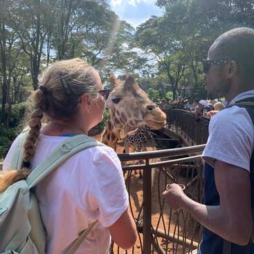 Two people stand by a fence, observing a giraffe at a wildlife park. The sunny day, trees, and other visitors create a lively, outdoor atmosphere.