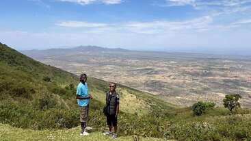 Two people stand on a green hillside, smiling, with a vast, sunlit valley and mountains stretching beneath a blue sky in the background. Serene landscape.