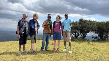 Five people stand on a grassy hilltop under a cloudy sky. They appear relaxed and happy, dressed for hiking, with scenic trees and landscape behind them.