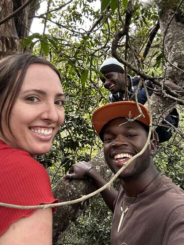 Three friends smile and pose for a selfie while climbing in a tree, surrounded by green leaves and branches, enjoying an adventurous and joyful outdoor moment together.