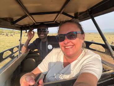 Two people smiling in a safari vehicle, wearing sunglasses. Open savanna landscape with grazing animals in the background, bright sunny day, joyful and adventurous atmosphere.