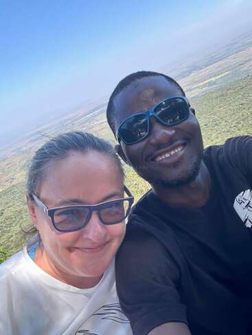 Un hombre y una mujer sonrientes con gafas de sol se toman un selfie al aire libre con un paisaje verde y un cielo azul de fondo. Parecen felices.