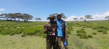 Dos hombres permanecen juntos al aire libre en una llanura cubierta de hierba con árboles dispersos bajo un cielo azul brillante. Uno lleva camuflaje, el otro ropa informal, ambos sonríen.