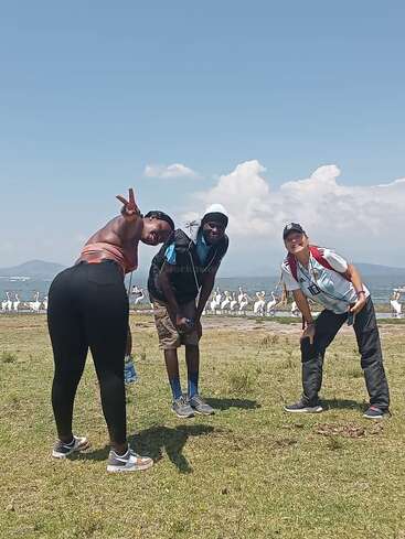 Tres amigos posan juguetones al aire libre cerca de un lago, con pelícanos al fondo. Es un día soleado, cielo azul, y todos parecen felices y relajados.