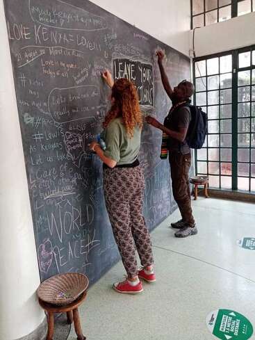 Dos personas escriben mensajes en una gran pizarra llena de palabras y dibujos positivos, bajo el lema "DEJA TUS ACCIONES", en un espacio luminoso y acogedor.