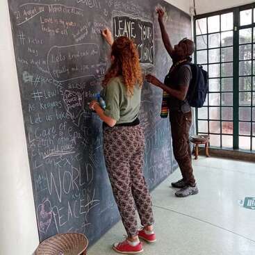 Two people write messages on a large chalkboard wall filled with positive words and drawings, under the prompt "LEAVE YOUR ACTIONS," in a bright, welcoming space.