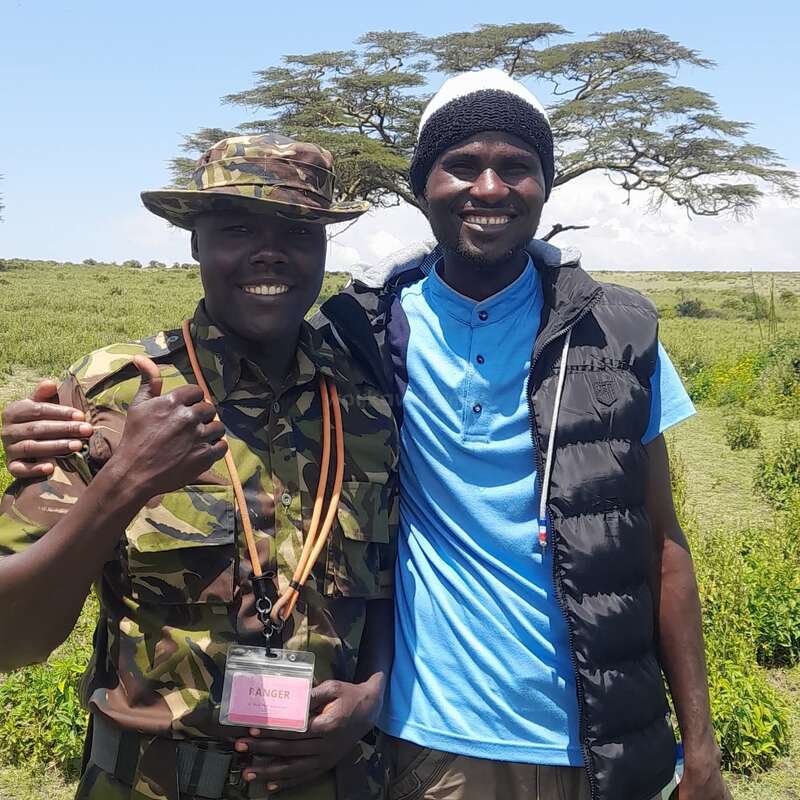Two smiling men stand together outdoors. One wears camouflage ranger uniform and hat, the other a blue shirt, black vest, and beanie. Green landscape and tree behind.