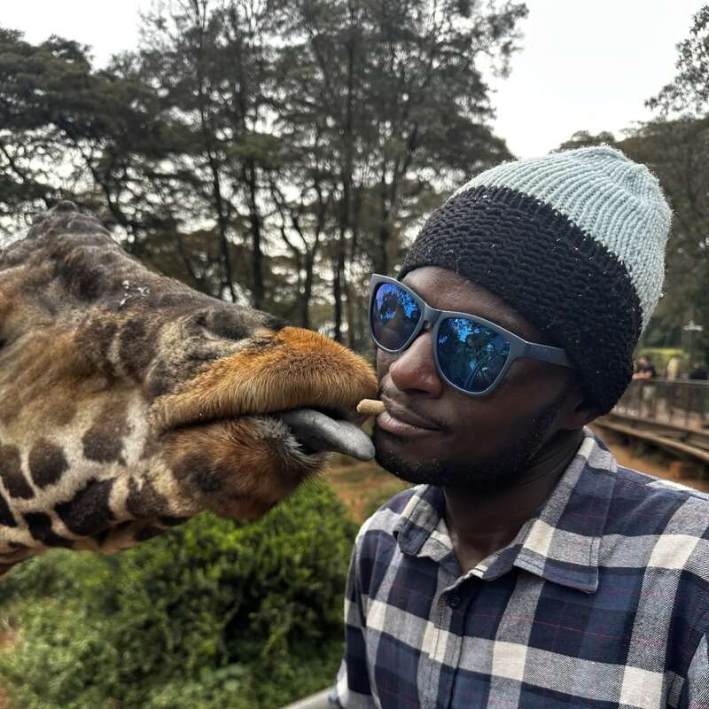 A man wearing sunglasses and a beanie is feeding a giraffe with his mouth, while the giraffe's tongue reaches out to grab the treat playfully.