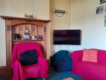 A cozy living room with a red-covered armchair and sofa, wooden mantelpiece decorated with animal figurines, wall-mounted TV, bean bag, and scatter cushions.