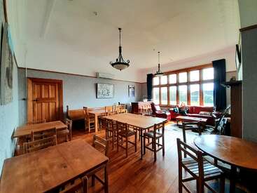 A spacious, well-lit room with wooden floors and furniture, large windows, red sofas, two chandeliers, and a person relaxing near natural light. Cozy atmosphere.