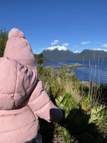Uma criança com uma jaqueta rosa e um chapéu olha para um lago tranquilo, cercado por plantas, com montanhas cobertas de neve incríveis sob um céu azul claro.