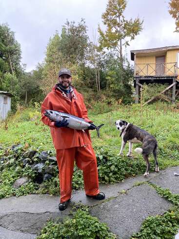 Um cara sorridente, com uma capa de chuva laranja, segura um peixe grande. Perto dele, um cachorro fica alerta. Árvores, grama e uma cabana amarela rústica completam a cena.