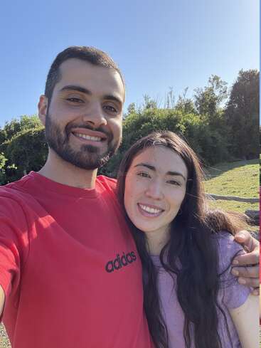 Um casal sorridente tira uma selfie ao ar livre em um dia ensolarado, bem juntinhos, com vegetação e céu azul ao fundo, curtindo a companhia um do outro.