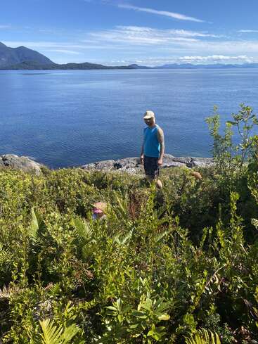 Un hombre con camisa azul y gorra está de pie en una costa rocosa, contemplando un lago tranquilo con montañas y un cielo azul al fondo, y una exuberante vegetación en primer plano.