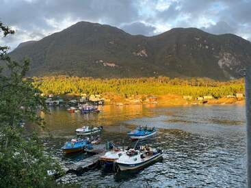 Vários barcos coloridos flutuam em um lago calmo, cercado por uma vegetação exuberante. Pequenas casas pontilham a costa ensolarada, enquanto grandes montanhas cobertas de florestas se erguem ao fundo.