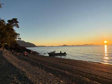 Uma cena tranquila na praia ao pôr do sol, com águas calmas, um pequeno barco flutuando, pessoas relaxando na praia, silhuetas de árvores, montanhas distantes e a luz dourada do sol refletindo lindamente.