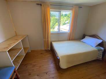 Este é um quarto aconchegante com piso de madeira, cama de solteiro, cortinas amarelas, janela com vista para a floresta, prateleiras e móveis simples e minimalistas.
