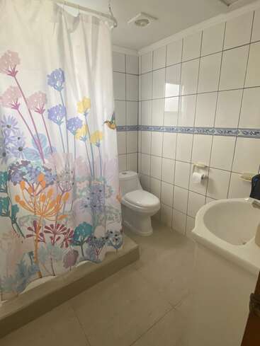 Esse banheiro tem paredes com azulejos brancos, um vaso sanitário, uma pia branca, piso bege e um chuveiro com uma cortina floral colorida pra dar um toque especial e garantir a privacidade.